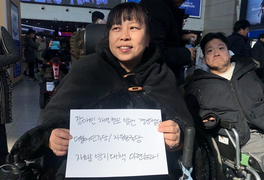 ‘장애인 차별험오 발언 경쟁정당 더불어민주당/자유한국당 재발방지대책 마련하라’는 문구가 쓰인 종이를 들고 있는 중증장애인 활동가.ⓒ전국장애인차별철폐연대 